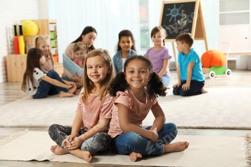 Preview: Cute girls sitting on floor while kindergarten teacher reading book to other children indoors