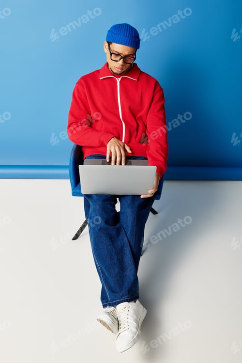 Preview: Stylish young african american man wearing a beanie on a bold blue backdrop with laptop
