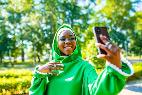 Preview: multicultural wwoman in green muslim dress with bright make-up and piercing nose taking selfie