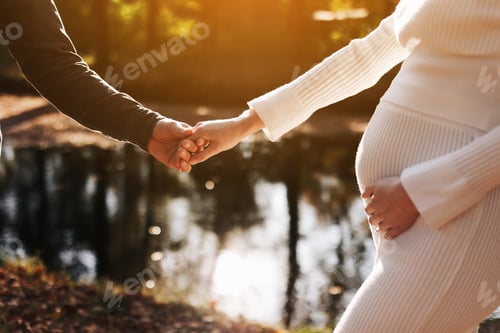 Preview: cropped photo of couple, man and pregnant wife are holding hands and walking by the lake on sun