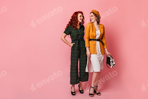 Preview: Full length view of young women in vintage outfit. Studio shot of cheerful friends talking on pink