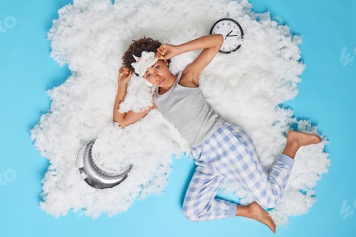 Preview: Sad young curly woman wears sleepmask and pajama has bad mood after seeing nightmare poses on white