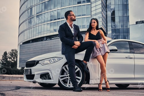 Preview: Well-dressed attractive couple leaning on a luxury car outdoors against the skyscraper.