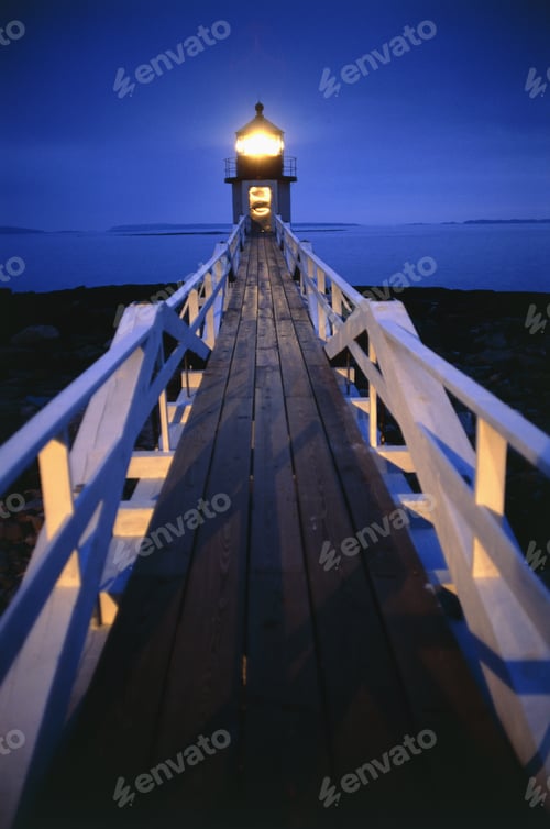 Preview: Illuminated wooden walkway towards lighthouse in the evening.