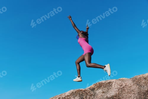 Preview: Woman jumping joyfully on a rock under a clear blue sky