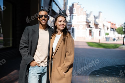Preview: happy and fashionable multiethnic couple in coats standing with hands in pockets on city street