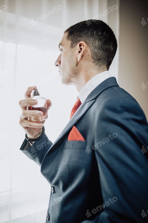 Preview: Vertical shot of a man in a suit looking through a window with a perfume in his hand