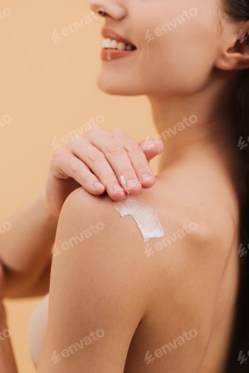 Preview: Smiling woman applying moisturizing cream on shoulder on a beige background.