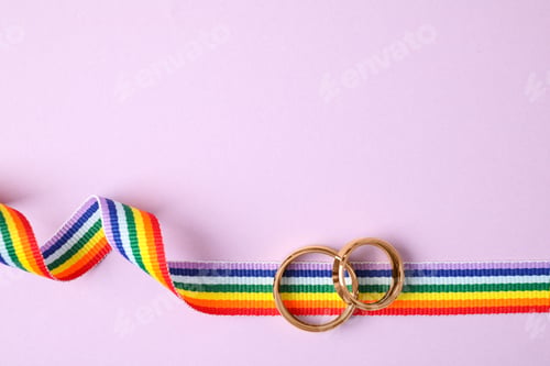 Preview: Top view of wedding rings and rainbow ribbon on color background, space for text. Gay symbol