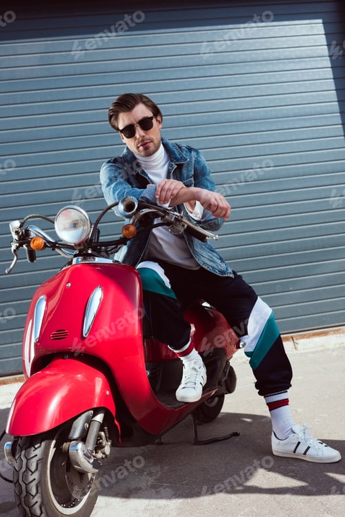 Preview: handsome young man in denim jacket and track pants sitting on vintage red scooter