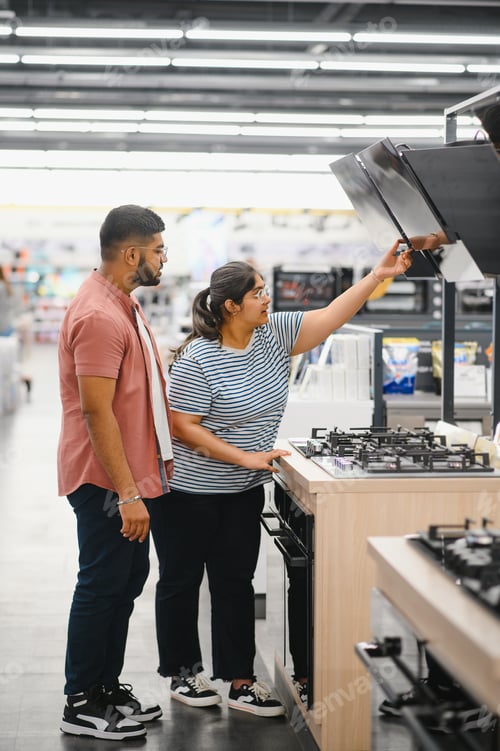 Preview: Couple of young customers looking at buildin cookers in domestic appliances shop