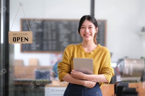 Preview: Happy young Asian woman owner standing holding a tablet with open sign board.