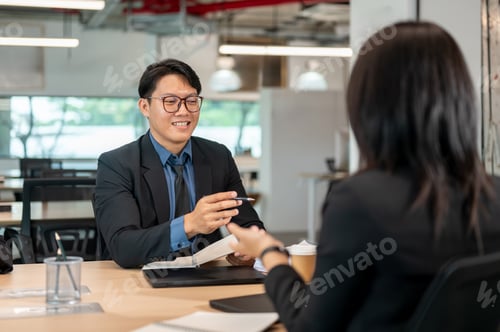 Preview: Businessman holding pen over notebook talking with woman coworker at wooden table in meeting room
