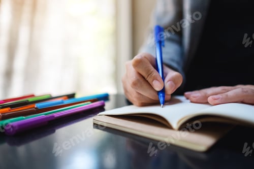 Preview: Closeup image of a woman writing on a blank notebook with colored pens on the table