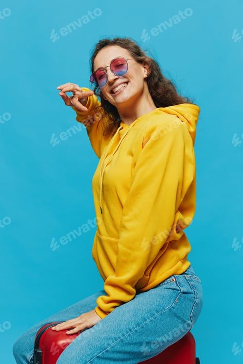 Preview: Smile woman sitting on a suitcase in a yellow hoodie, blue jeans and glasses on a blue background