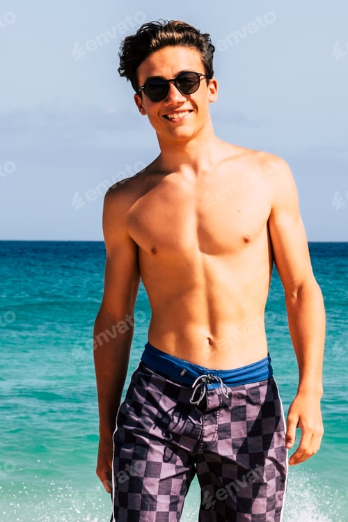 Preview: Young handsome teenager in swimsuit walking at the beach with blue sky and ocean water