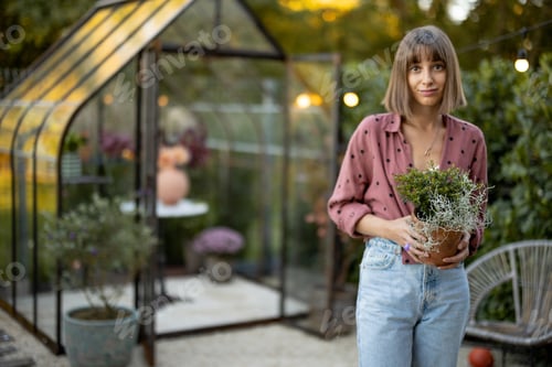 Preview: Woman with flowerpot in garden with greenhouse behind