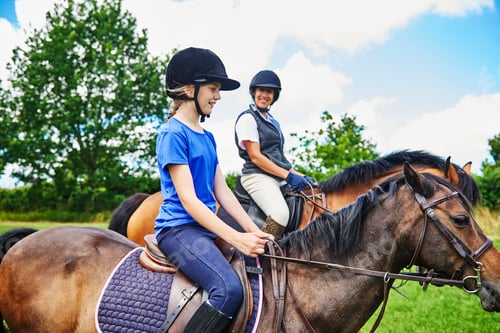 Preview: Side view of mature woman and girl on horseback wearing riding hats smiling