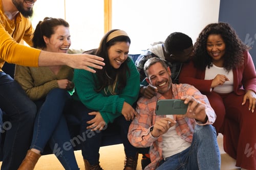 Preview: Happy diverse male and female friends relaxing at home together talking and looking at smartphone