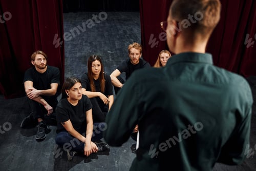 Preview: Sitting and listening to man. Group of actors in dark colored clothes on rehearsal in the theater