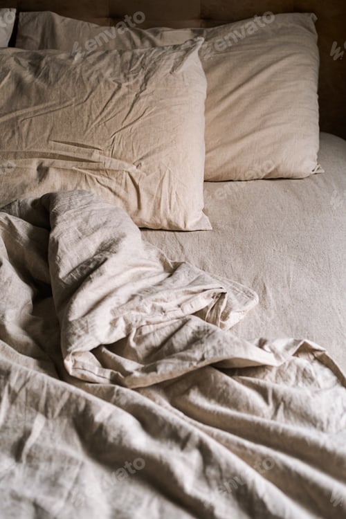 Preview: Empty bed covered wrinkled bedding and pillows in cozy bedroom at nighttime.