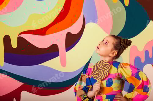 Preview: Stylish little girl posing with round large lollipop at multi colours wall backdrop