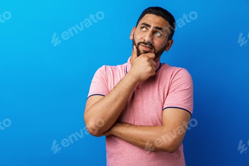 Preview: Pensive young arab man thinking and looking up against blue background