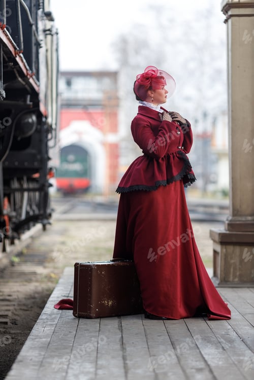 Preview: A girl in a burgundy suit and a hat with a veil stands with a suitcase near an old steam locomotive