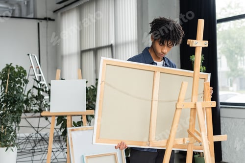 Preview: Young african american artist holding canvas near wooden easel in studio