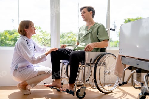 Preview: Senior Caucasian doctor woman sit and talk to young patient man who sit on wheelchair in room