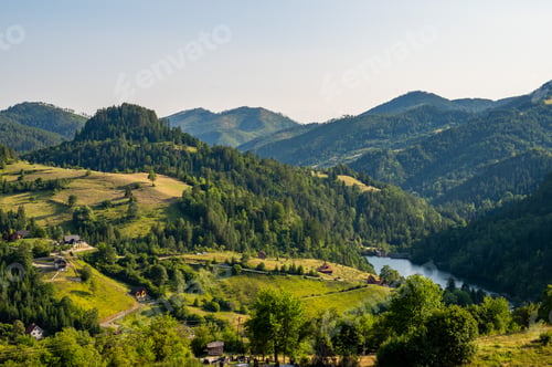 Preview: Lush green valley with rural houses surrounded by forested mountains under clear blue sky.