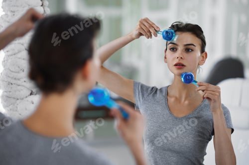 Preview: A young woman is massaging her face with blue glass balls while looking in the mirror.