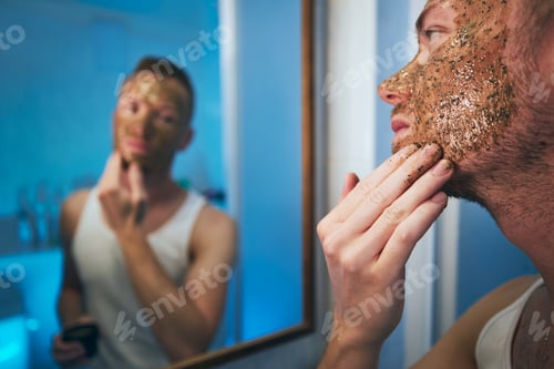 Preview: Young man applying facial mask in front of mirror in home bathroom