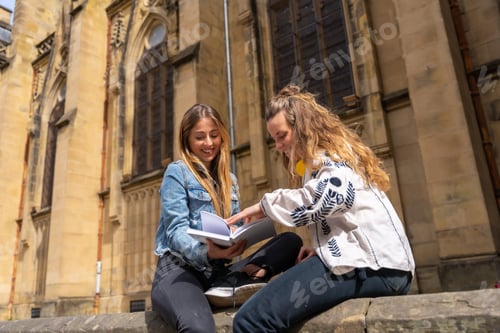 Preview: Two young Caucasian women in the city reading a book sitting next to a church