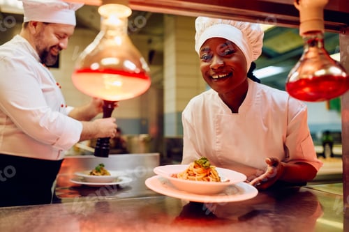 Preview: Happy black female chef serving a dish in restaurant kitchen and looking at camera.