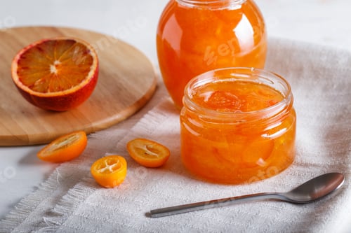 Preview: Orange and kumquat jam in a glass jar with fresh fruits on a white linen tablecloth.