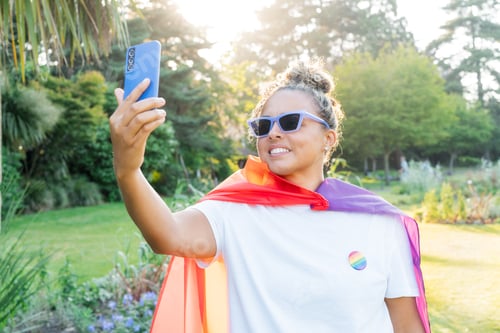 Preview: Young Latin happy woman wrapped in a rainbow flag, symbol of LGBT community, makes selfie on phone