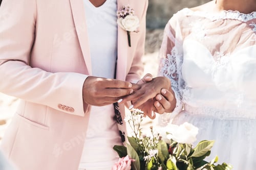 Preview: Groom in pink jacket wears ring on the finger of the bride on the beach