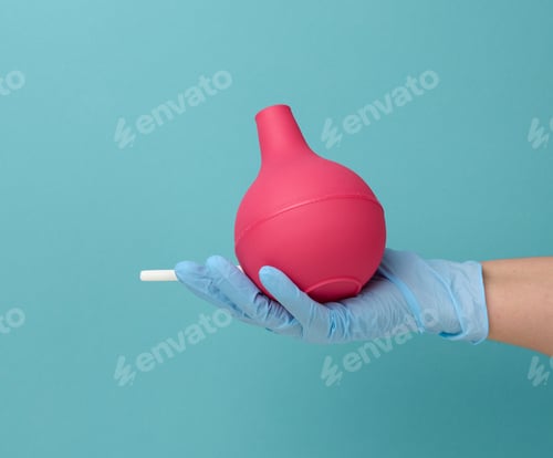 Preview: Hand in a blue medical glove holds a pink rubber enema on a blue background