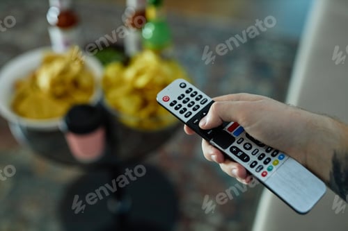 Preview: Hand Holding TV Remote Choosing Channels with Snacks Bottles in Background