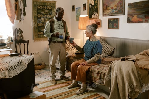 Preview: Senior Caucasian Woman and Senior Black Man Sharing Hot Drinks in Cozy Living Room