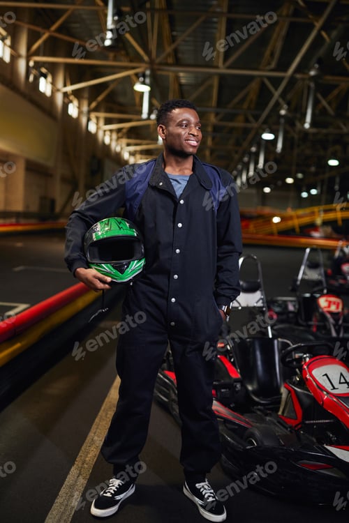 Preview: happy african american man in sportswear holding helmet and standing near go-kart in karting track
