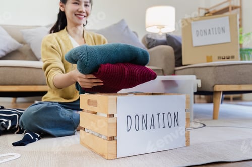 Preview: Young Asian women sit in living room sorting clothes for donation in a donation box second hand