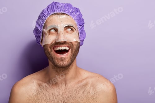 Preview: Happy positive young man applies facial mask, wears shower cap, laughs positively, has naked body, d