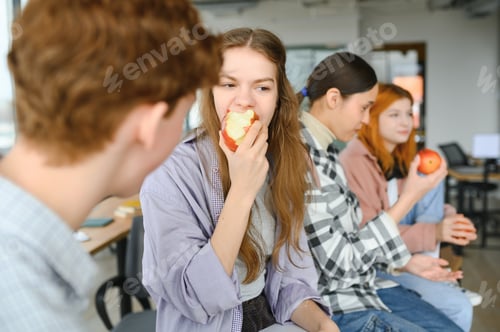 Preview: Cheerful students are sitting at desks and eating an apple during a break