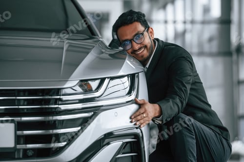Preview: Embracing the transport. Handsome indian man in suit is in the car showroom with the vehicle indoors