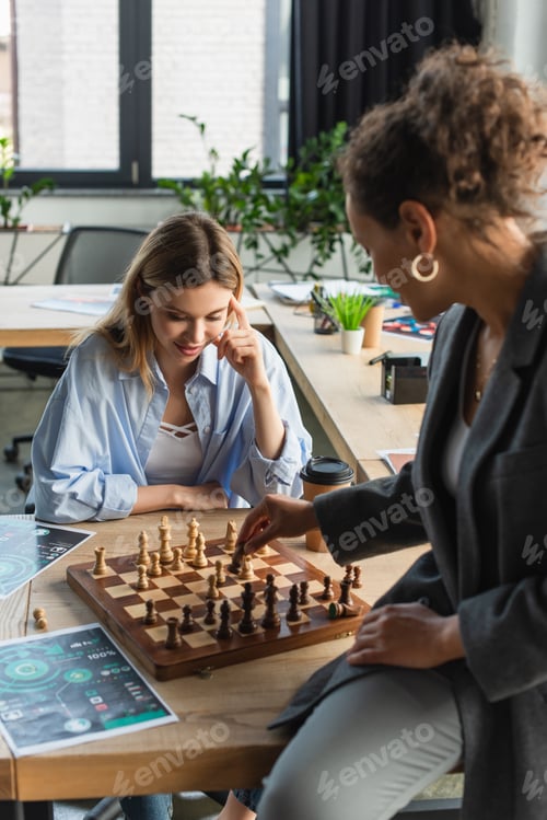 Preview: Pensive businesswoman looking at chess on board while playing with african american colleague in