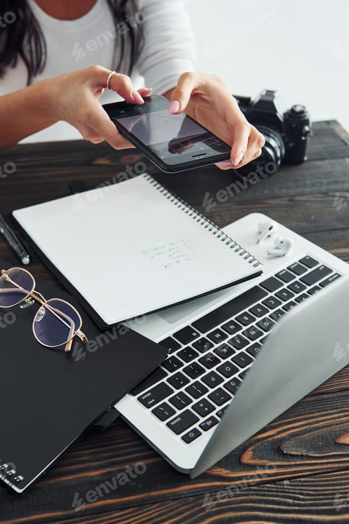 Preview: Holding phone. Young female freelancer working indoors in the office at daytime. Laptop and notepad