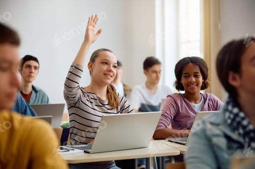 Preview: Teenage girl using laptop and raising her hand to ask question during computer class at high school.