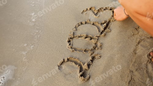 Preview: Closeup of young woman sitting at sea beach and writing 2023 year on sand